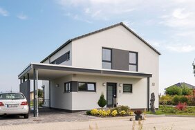 Außenansicht des Finger-Haus Musterhauses MEDLEY 300 B in Berlin/Werder: zweigeschossiges Haus mit weißer Fassade und grauen Akzentflächen, Satteldach und Carport; davor gepflasterte Einfahrt mit geparktem Auto, Beetbepflanzung, Rasen und Bäume unter blauem Himmel.