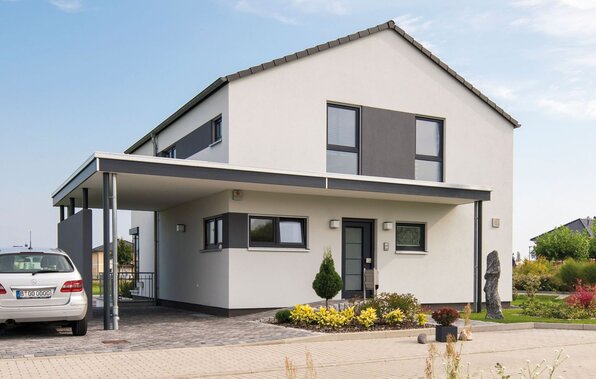 Außenansicht des Finger-Haus Musterhauses MEDLEY 300 B in Berlin/Werder: zweigeschossiges Haus mit weißer Fassade und grauen Akzentflächen, Satteldach und Carport; davor gepflasterte Einfahrt mit geparktem Auto, Beetbepflanzung, Rasen und Bäume unter blauem Himmel.