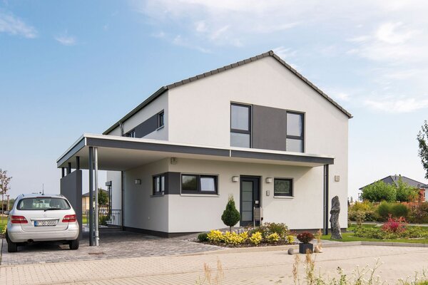 Außenansicht des Finger-Haus Musterhauses MEDLEY 300 B in Berlin/Werder: zweigeschossiges Haus mit weißer Fassade und grauen Akzentflächen, Satteldach und Carport; davor gepflasterte Einfahrt mit geparktem Auto, Beetbepflanzung, Rasen und Bäume unter blauem Himmel.