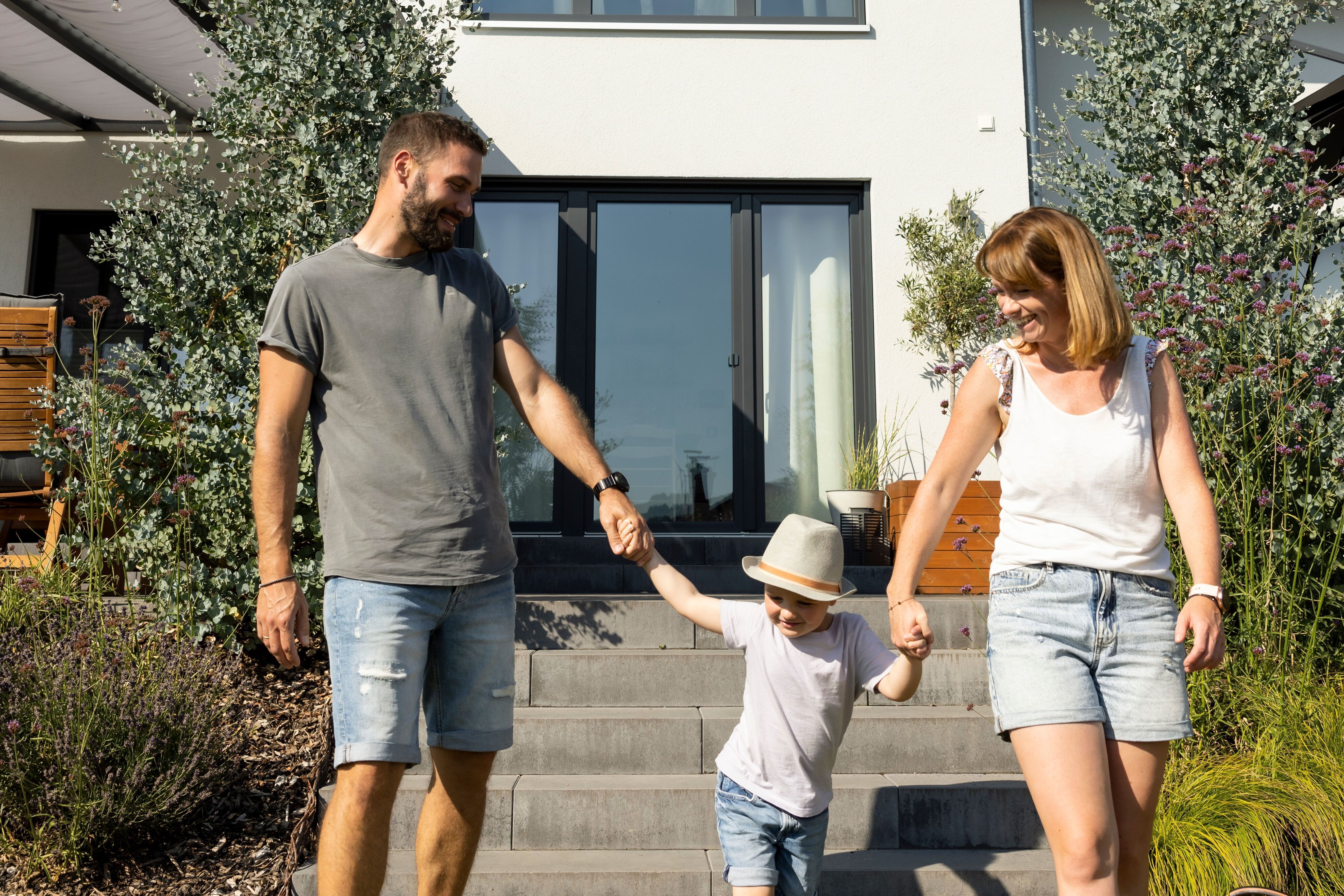 Familie mit zwei Erwachsenen und einem Kind läuft Hand in Hand über den Garten vor einem Einfamilienhaus mit Satteldach