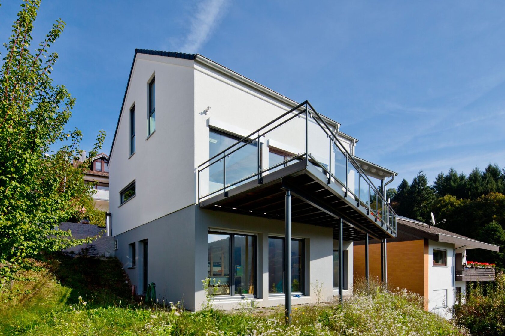 Frei geplantes Haus von FingerHaus mit zweigeschossiger Architektur bei Tageslicht, helle Putzfassade mit dunklem Sockelbereich, großzügiger Balkon mit Glasgeländer, bodentiefe Fenster und Hanglage mit Grünfläche.