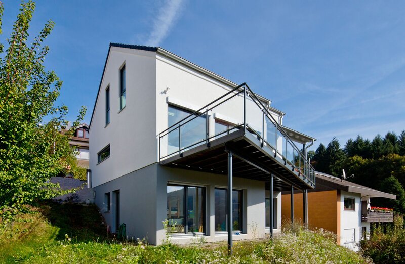Frei geplantes Haus von FingerHaus mit zweigeschossiger Architektur bei Tageslicht, helle Putzfassade mit dunklem Sockelbereich, großzügiger Balkon mit Glasgeländer, bodentiefe Fenster und Hanglage mit Grünfläche.