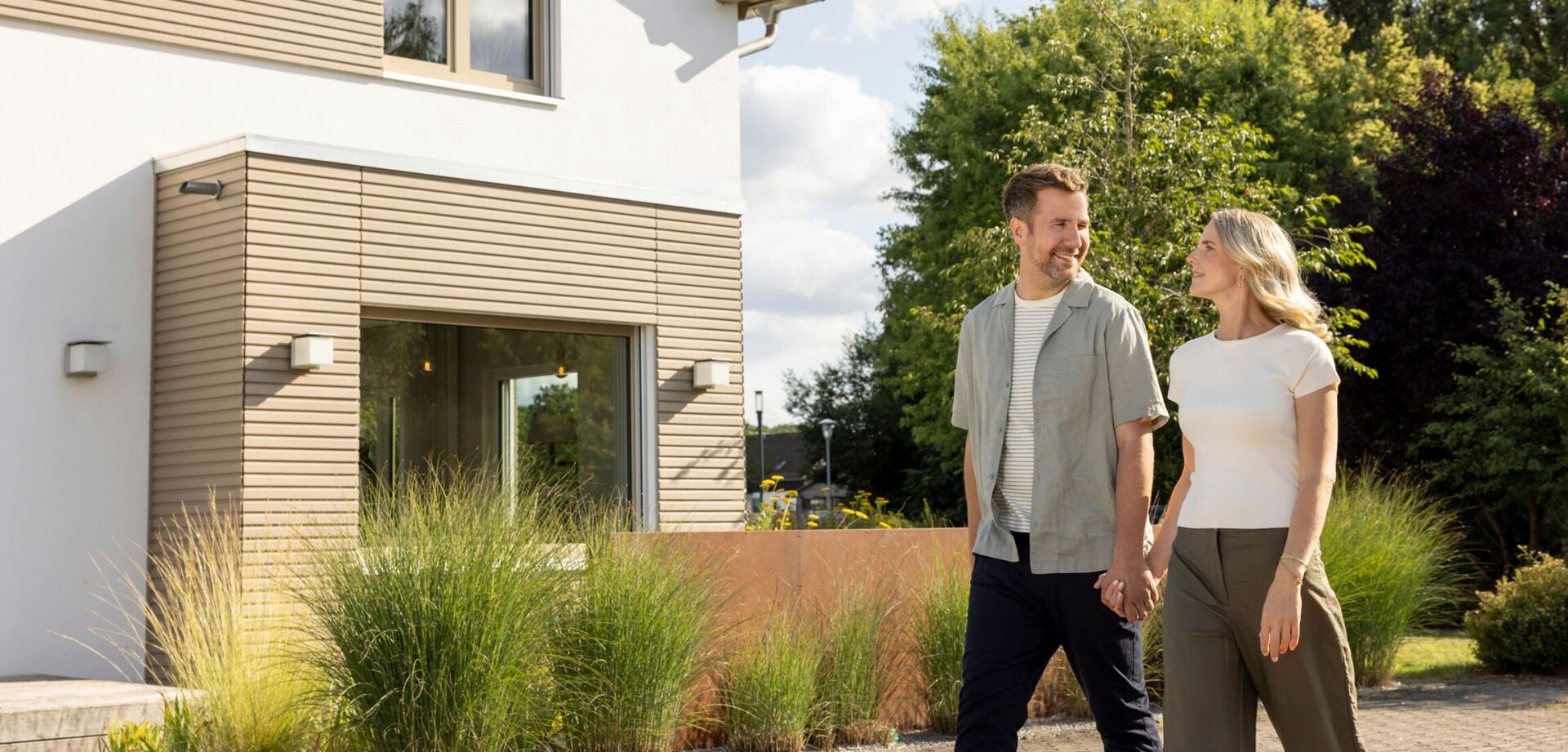 Paar geht Hand in Hand durch den Musterhauspark von FingerHaus in Frankenberg, im Vordergrund ein modernes zweigeschossiges Haus mit heller Putzfassade, Holzverkleidung und großen Fenstern, umgeben von Grünflächen.