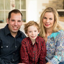 Eine Familie sitzt gemeinsam auf dem Sofa und lächelt in die Kamera. Im Hintergrund ist ein modern eingerichtetes Wohnzimmer zu sehen.