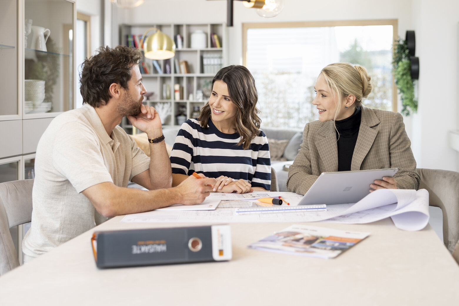 Drei Personen sitzen an einem Tisch mit Bauplänen und Ordnern in einem hellen, modernen Raum und besprechen etwas.