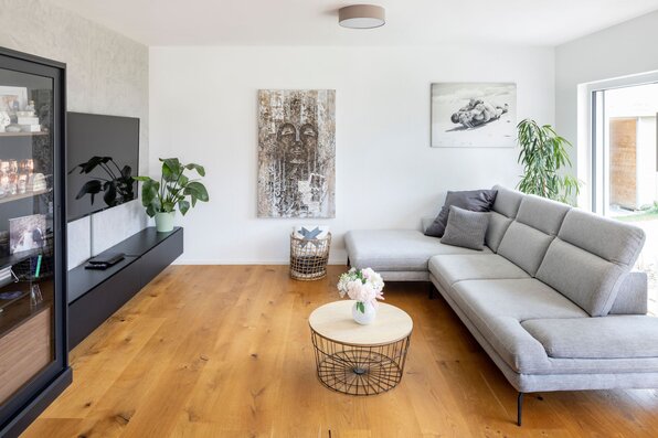 Wohnzimmer mit grauem Ecksofa, rundem Couchtisch, wandmontiertem Fernseher, Holzfußboden und mehreren Zimmerpflanzen.