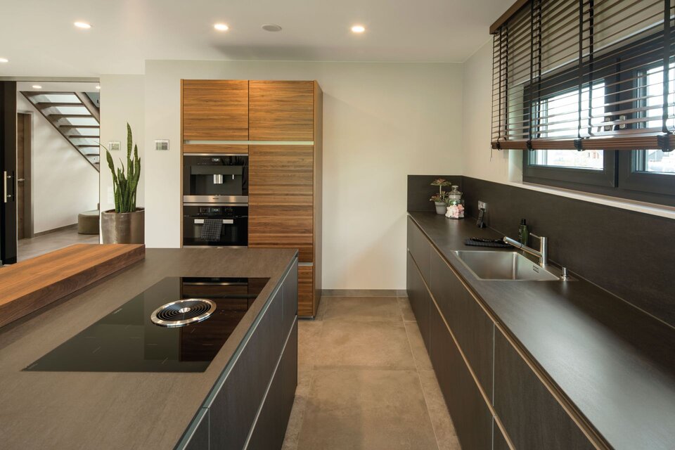 Moderne Küche mit Kochinsel, schwarzen Arbeitsflächen und Holzschränken, rechts Fenster mit Jalousien und links Treppe mit Pflanzen