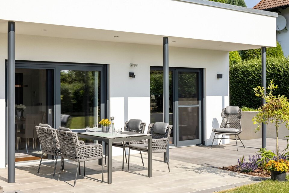 Überdachte Terrasse eines frei geplanten FingerHaus im modernen Bauhaus-Stil mit Sitzgruppe aus geflochtenen Stühlen und Tisch. Bodentiefe Fenster führen in den Wohnbereich, daneben gepflegte Bepflanzung.