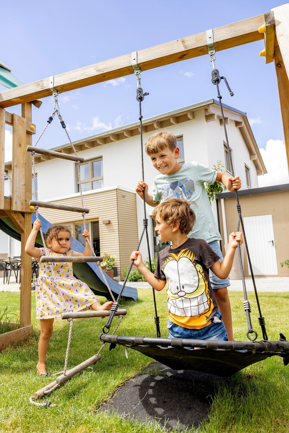 Drei Kinder spielen auf zwei Schaukeln im Garten vor einem weißen Einfamilienhaus.