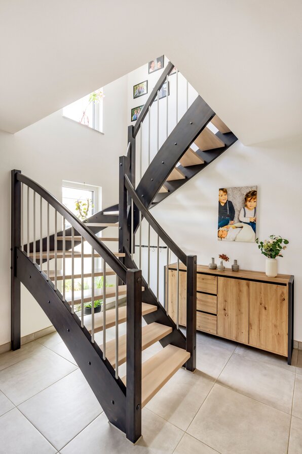 Offenes Treppenhaus mit dunkler Holztreppe, hellen Stufen und Metallstäben im Geländer sowie einer Holzkommode an der Wand.