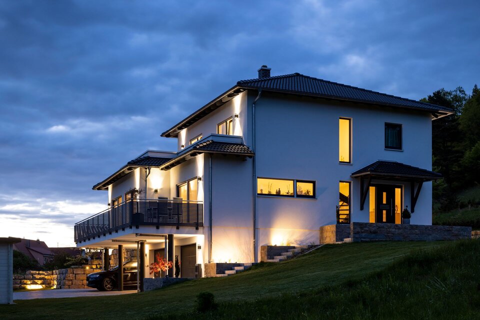 Beleuchtetes modernes FingerHaus mit Walmdach in der Abenddämmerung, großes Wohnhaus mit Balkon und Garage.