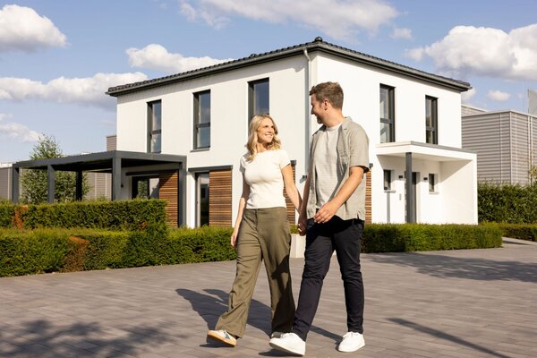 Paar spaziert im Musterhauspark Frankenberg vor dem FingerHaus SENTO mit weißer Fassade und Walmdach.