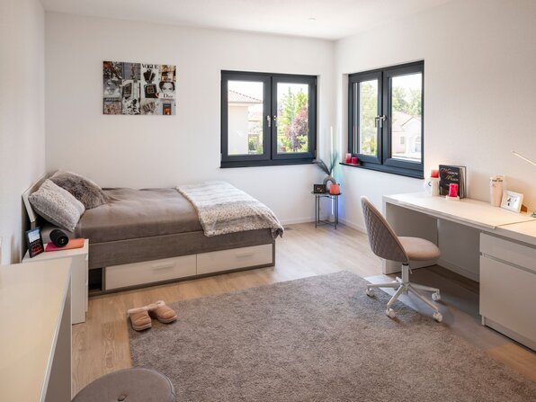 Schlafzimmer mit Bett, Schreibtisch, Stuhl, Teppich und Eckfenstern.