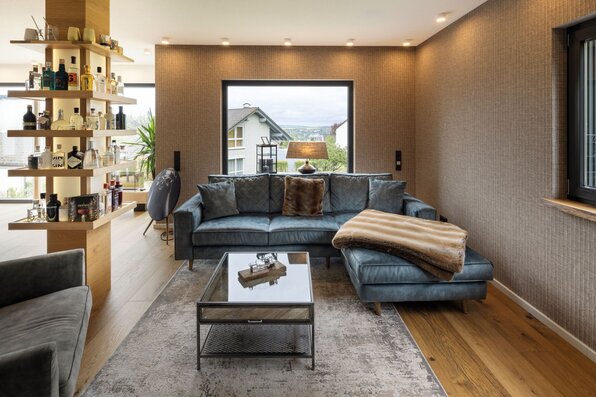 Gemütliches Wohnzimmer in einem modernen FingerHaus-Haus mit großer Couch in Blautönen, Couchtisch aus Glas und Metall sowie stilvoller Bar-Regalwand aus Holz.