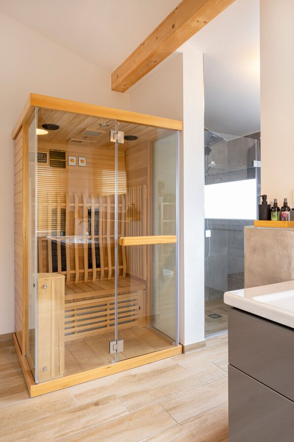 Badezimmer mit verglaster Holzsauna neben einer bodengleichen Dusche sowie einem grauen Waschtischunterschrank.