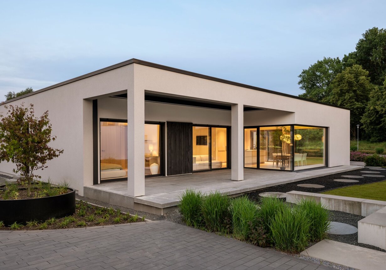 Außenansicht eines Bungalow NIVO mit Flachdach, überdachter Terrasse, großen Fensterfronten und modern gestalteter Gartenanlage.