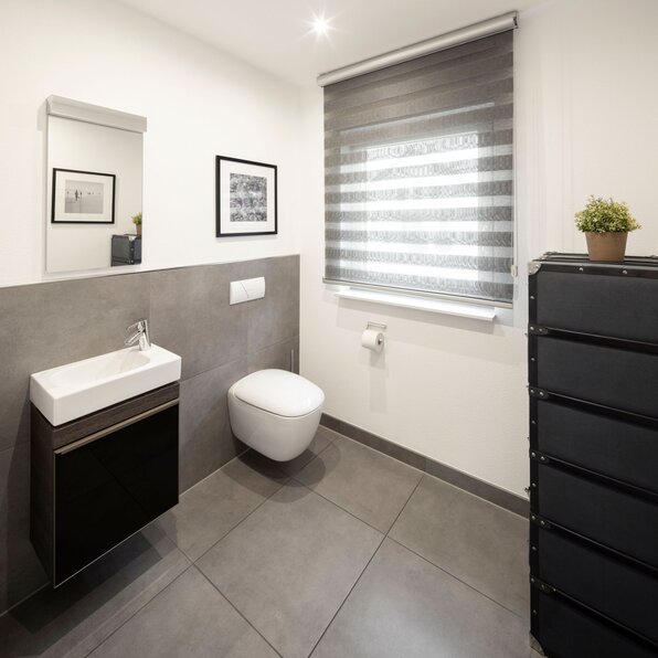 Modernes Gäste-WC im Musterhaus NEO 312 in Bad Vilbel mit wandhängendem WC, kleinem Waschbecken und stilvollem grauen Fliesen-Design.