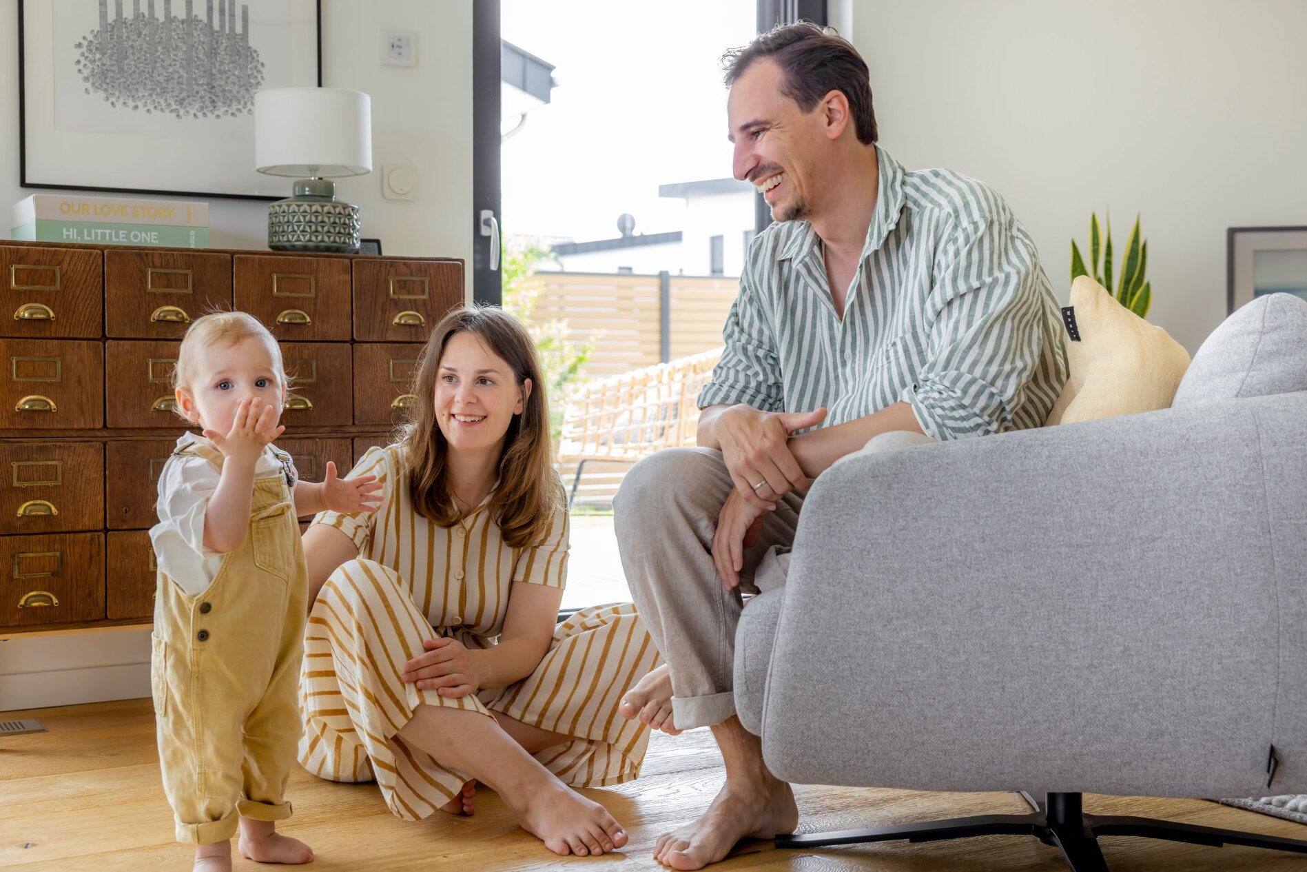 Eine junge Familie sitzt barfuß im Wohnzimmer ihres modernen FingerHaus-Hauses. Das Kind steht neugierig im Vordergrund, während die Eltern lachend zusehen.