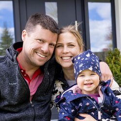 Ein Mann, eine Frau und ein kleines Kind sitzen lachend auf einer Steinstufe vor einem modernen Einfamilienhaus. Das Kind trägt eine blaue Jacke mit Kapuze und Mütze, die Erwachsenen sind leger gekleidet.