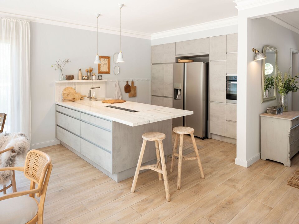 Moderne Küche mit großer Kochinsel in Betonoptik, hellen Fronten, zwei Holzhockern, offenen Regalen und Holzfußboden.