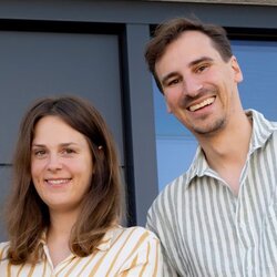 Eine junge Familie steht lächelnd vor der Eingangstür ihres modernen FingerHaus-Hauses. Die Mutter hält das Baby im Arm, während der Vater daneben steht.