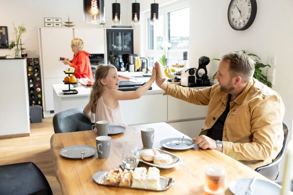 Drei Generationen verbringen Zeit im offenen Wohn- und Essbereich des Kundenhauses NIVO 80. Ein Mann und ein Kind geben sich am Esstisch ein High Five, während eine Seniorin in der Küche steht.
