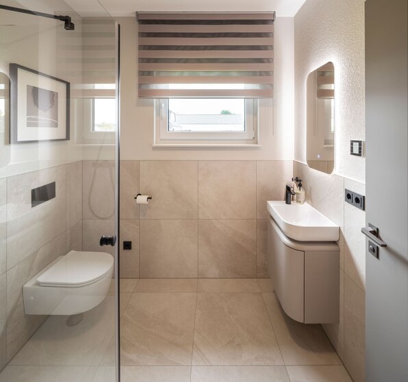 Modernes Badezimmer im Musterhaus JUNO 300 in Mülheim-Kärlich mit bodengleicher Dusche, wandhängendem WC, Waschbecken mit LED-Spiegel und großem Fenster.