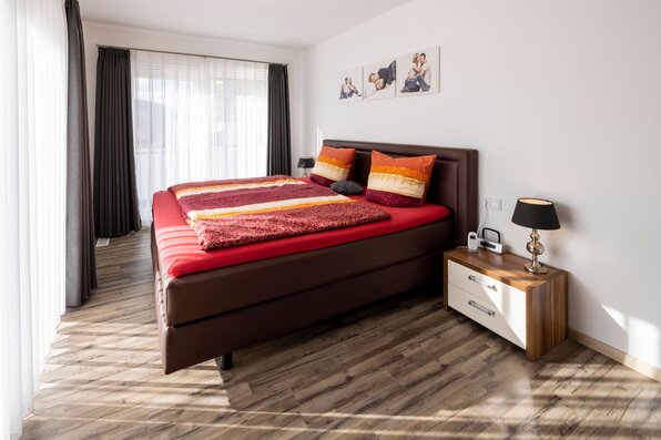 Schlafzimmer mit Doppelbett, zwei bunten Kissen und Decken, Nachttisch mit Lampe und Telefon, helle Vorhänge und Holzboden