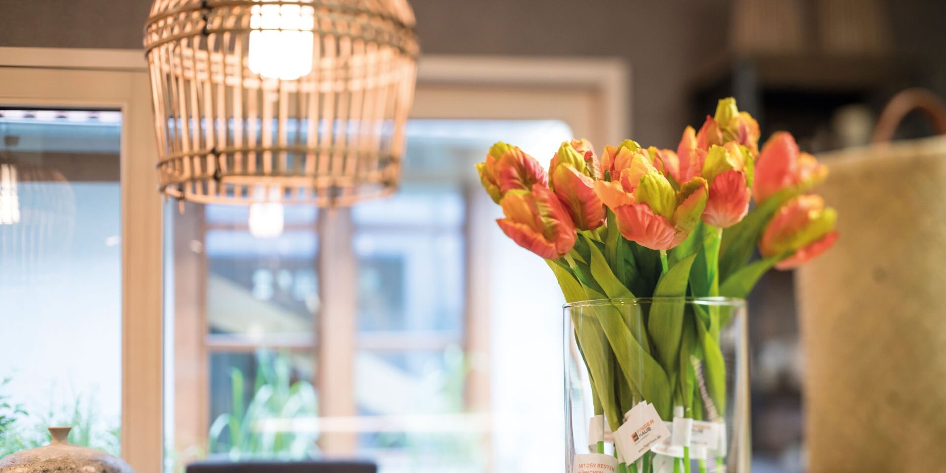 Glasvase mit orangefarbenen Tulpen auf einem weißen Tisch in einem modernen Innenraum mit Hängeleuchte und Fenster im Hintergrund