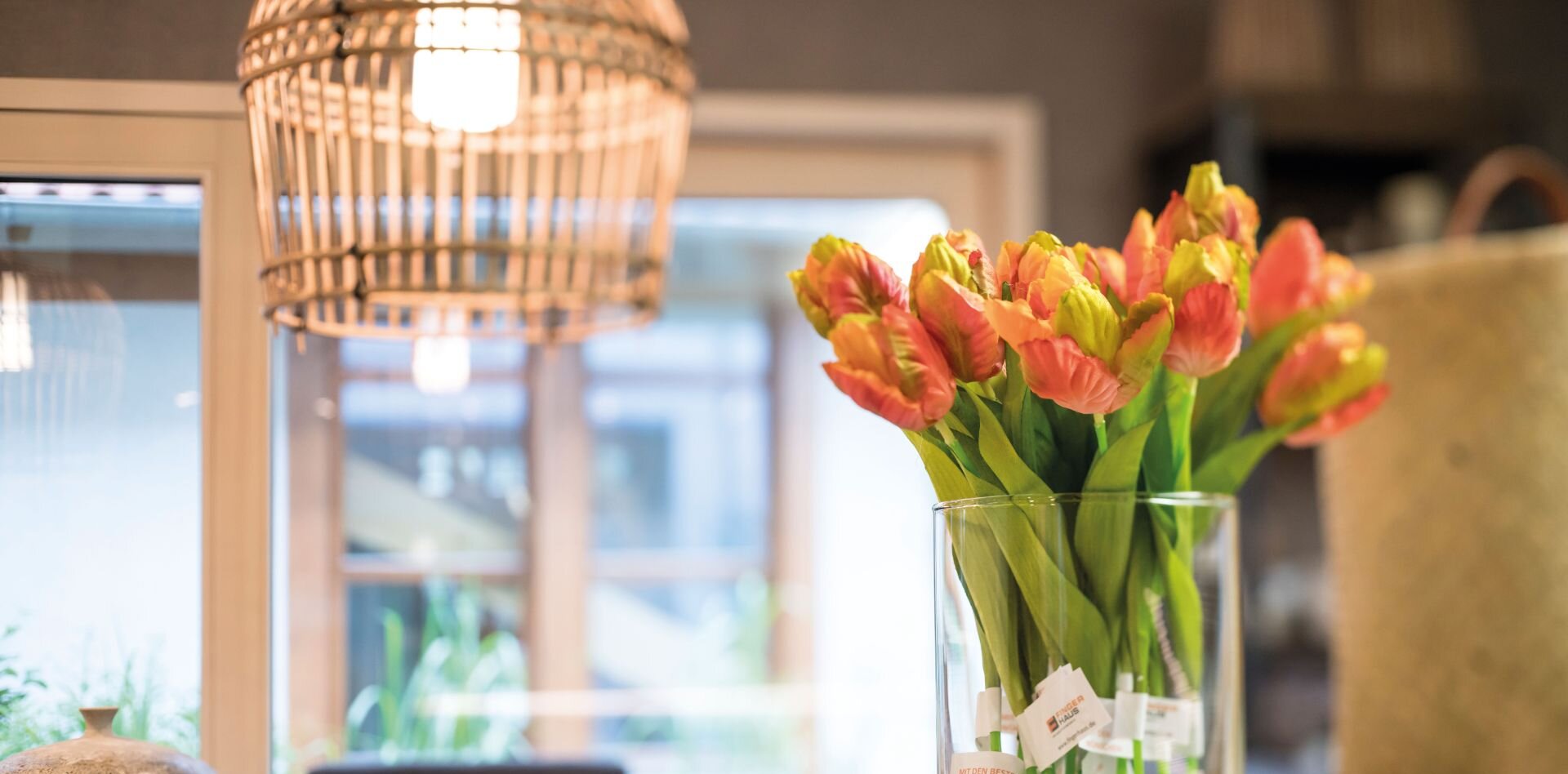 Glasvase mit orangefarbenen Tulpen auf einem weißen Tisch in einem modernen Innenraum mit Hängeleuchte und Fenster im Hintergrund