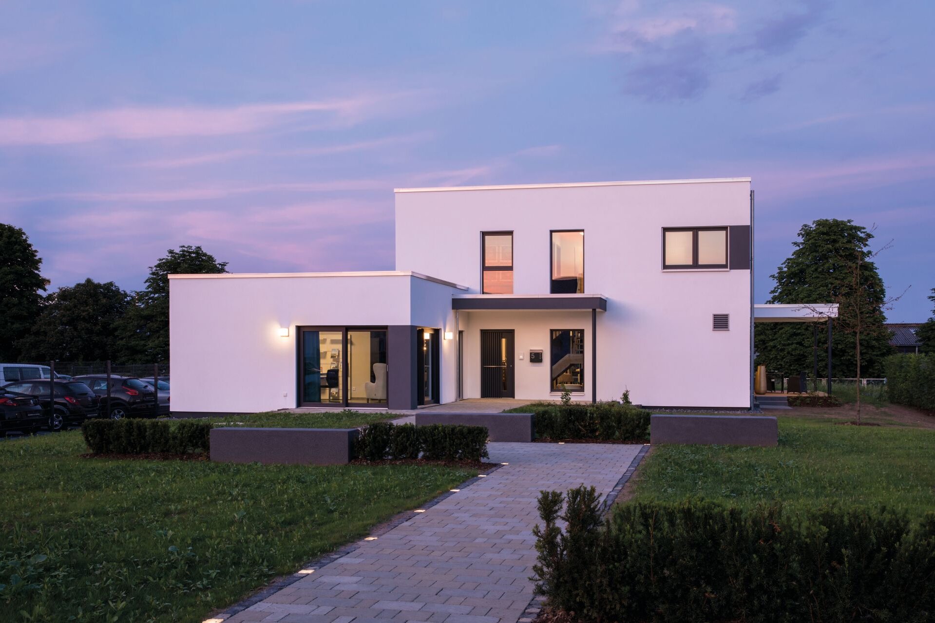 Musterhaus ALTERO in Gießen mit moderner Flachdacharchitektur, weißer Putzfassade, beleuchtetem Eingangsbereich und gepflastertem Zugangsweg in der Abenddämmerung.