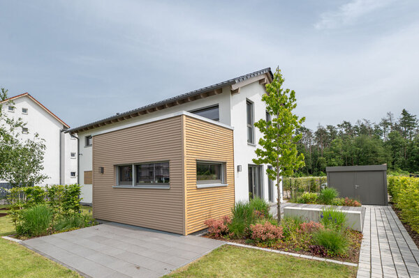 Außenansicht des Finger-Haus Musterhauses TALO in Babenhausen mit weißer Putzfassade und großem Holzfassaden-Anbau, Rasenfläche mit Trittplatten, gepflasterter Terrasse, bepflanzten Beeten, Hecken und gepflastertem Weg zu einem grauen Gartenhaus.