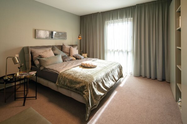 Gemütliches Schlafzimmer im Musterhaus MEDLEY 300 B in Frankenberg mit großen Vorhängen, Doppelbett und modernen Nachttischen.