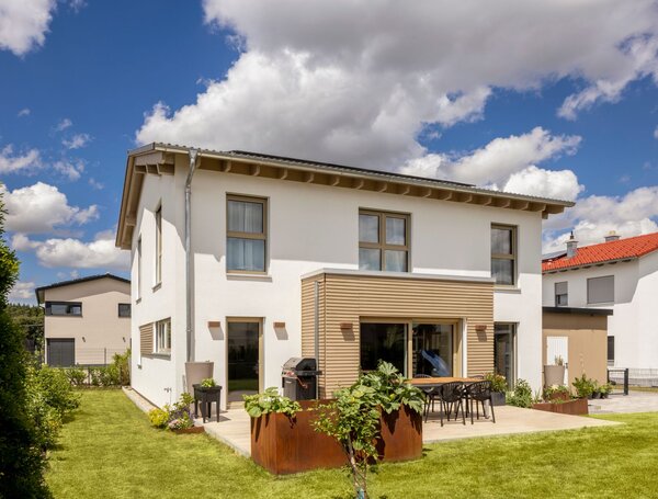 Zweigeschossiges weißes Einfamilienhaus mit flach geneigtem Dach, großen Fenstern, Holzverkleidung und Terrasse mit Esstisch im Garten.