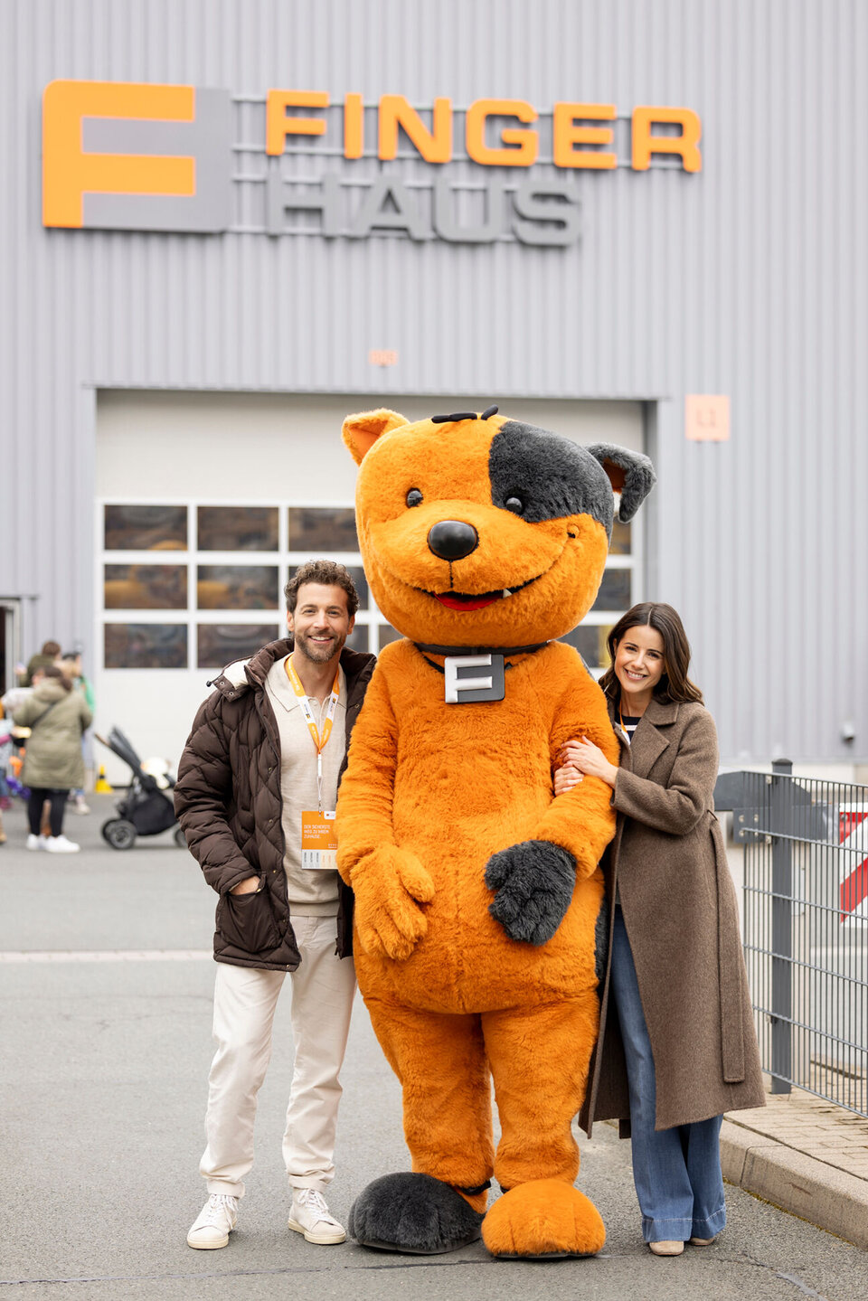 Ein Paar steht beim FingerHaus-Bauinfotag in Frankenberg neben dem orange-schwarzen Maskottchen FRÄNKIE. Im Hintergrund ist eine Halle mit dem FingerHaus-Logo und weiteren Besuchern zu sehen.