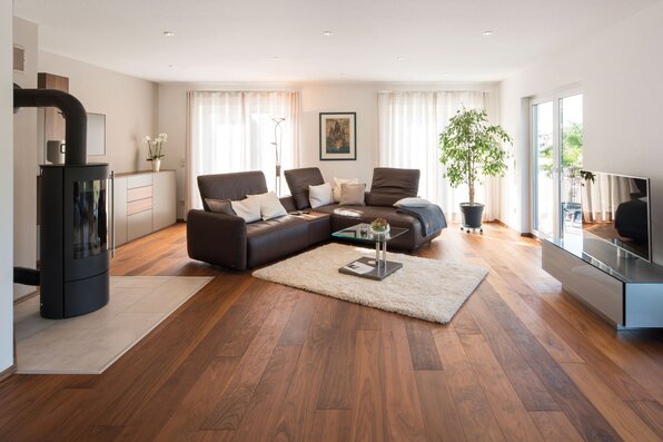 Helles Wohnzimmer in einem frei geplanten FingerHaus mit brauner Ledercouch, Kaminofen, Glastisch, TV-Bereich und großen Fenstern mit Blick in den Garten.