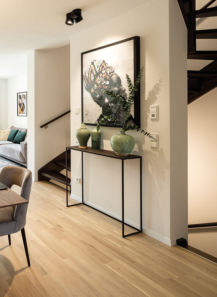Modern eingerichteter Essbereich mit Holztisch, sechs gepolsterten Stühlen und großer runder Hängelampe; im Hintergrund offener Wohnbereich und Treppe.