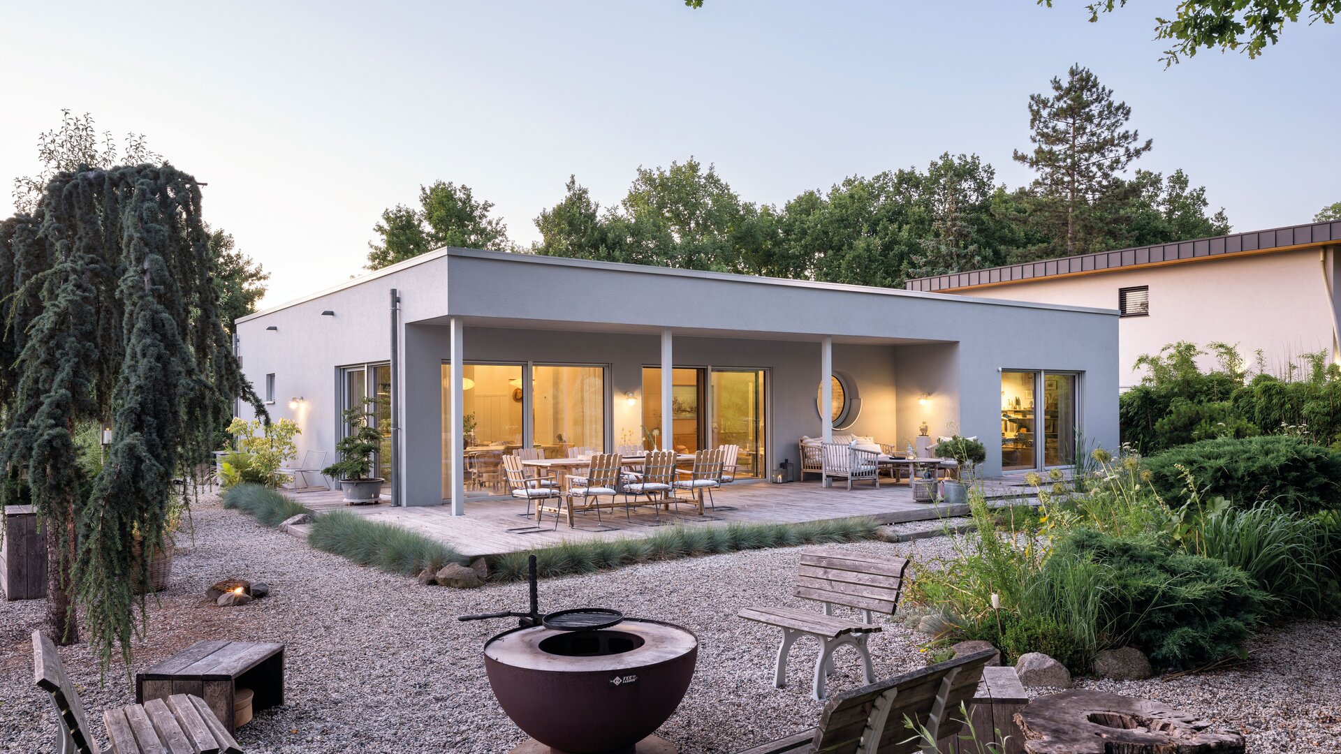 Außenansicht eines modernen eingeschossigen Hauses mit Flachdach, breiten Fensterfronten, überdachter Terrasse, Gartenmöbeln, Feuerstelle und Kiesgarten im Vordergrund.
