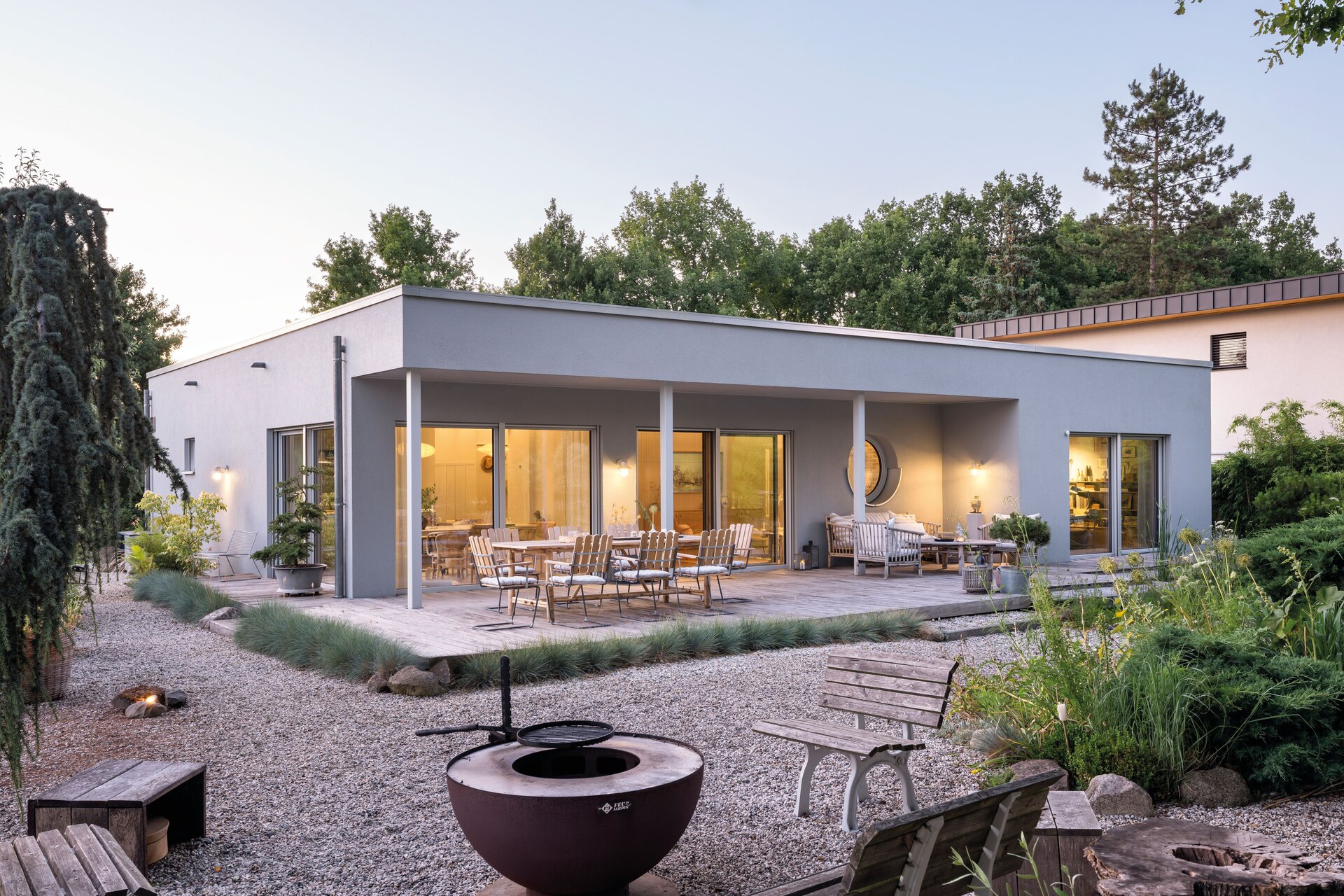 Außenansicht eines modernen eingeschossigen Hauses mit Flachdach, breiten Fensterfronten, überdachter Terrasse, Gartenmöbeln, Feuerstelle und Kiesgarten im Vordergrund.