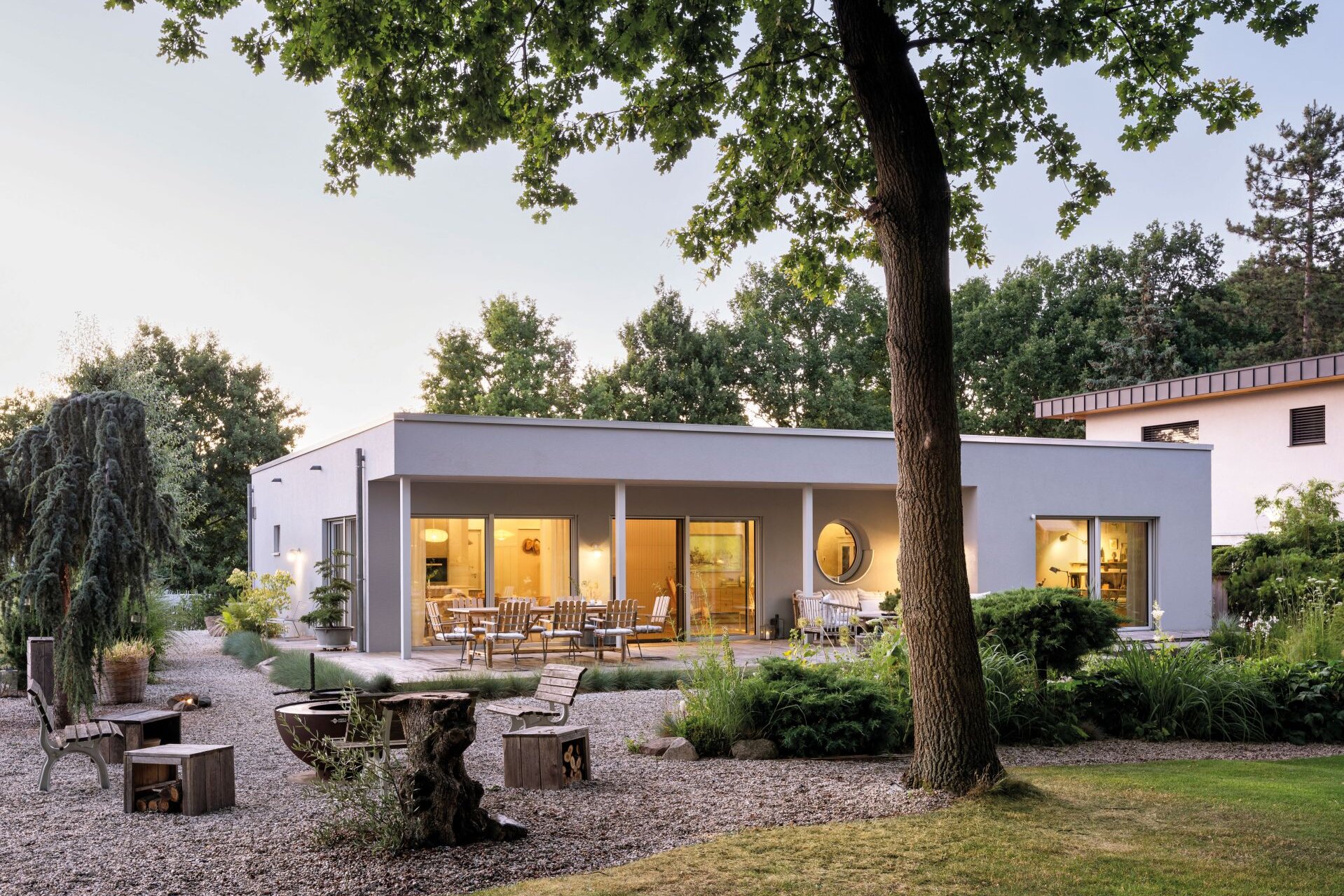 Flachdachhaus mit großer Fensterfront und überdachter Terrasse in einem Garten mit Bäumen und Sitzbereich