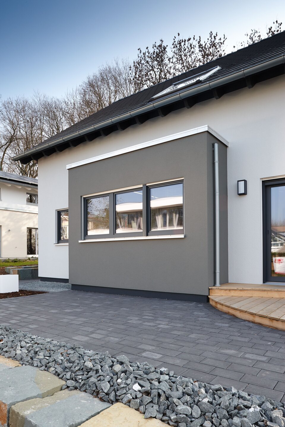 Seitenansicht eines modernen Musterhauses mit grauem Erker und dunklem Satteldach. Vor dem Haus liegen Pflastersteine und eine Begrenzung aus Naturstein und Zierkies.