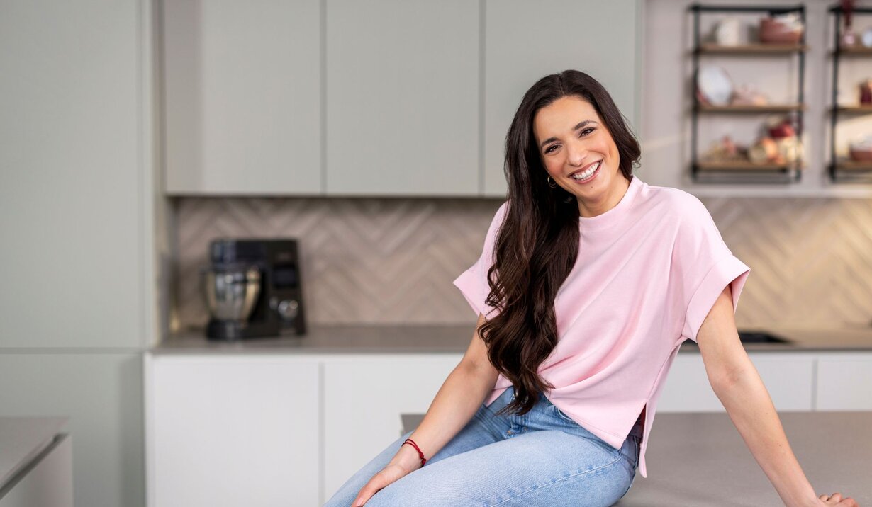 Sally Özcan, bekannte Influencerin, mit langen dunklen Haaren sitzt auf einer Küchenarbeitsplatte, trägt ein helles T-Shirt und blaue Jeans, im Hintergrund moderne Küchenmöbel und ein Kaffeevollautomat.