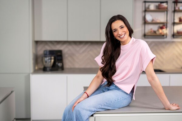 Sally Özcan, bekannte Influencerin, mit langen dunklen Haaren sitzt auf einer Küchenarbeitsplatte, trägt ein helles T-Shirt und blaue Jeans, im Hintergrund moderne Küchenmöbel und ein Kaffeevollautomat.