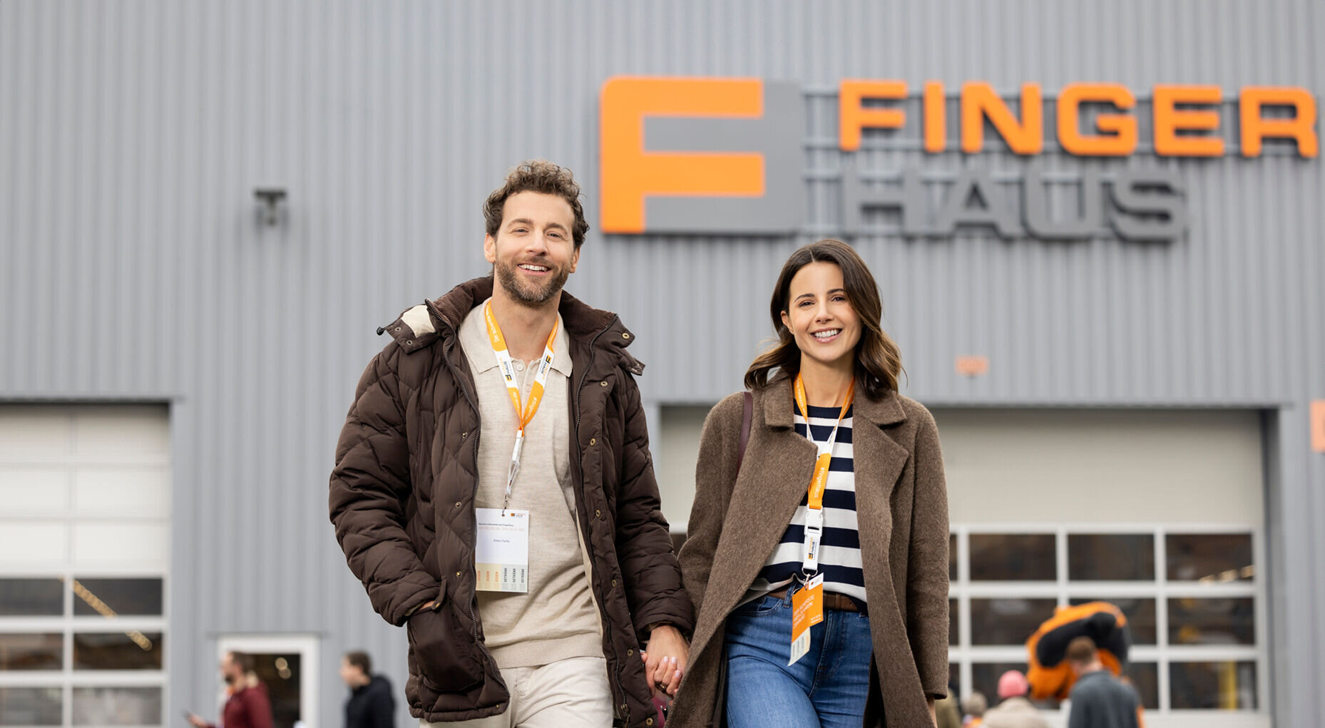 Ein Paar geht lächelnd über das Veranstaltungsgelände des FingerHaus-Bauinfotags in Frankenberg. Im Hintergrund ist eine Halle mit dem FingerHaus-Logo zu sehen, umgeben von weiteren Besuchern.