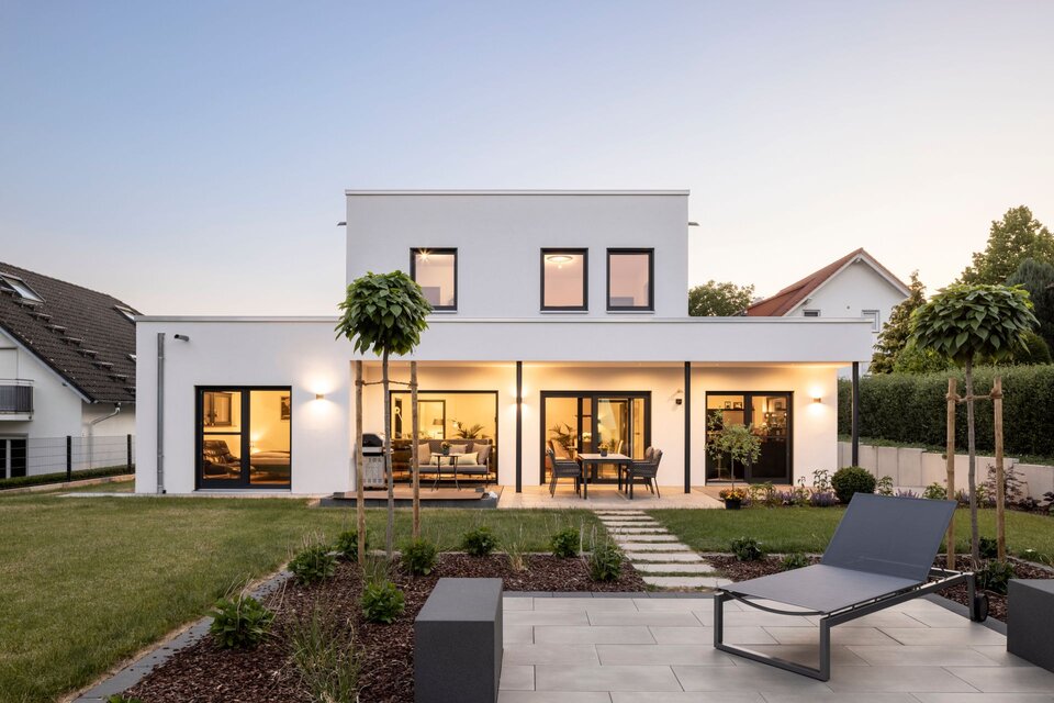 Abendliche Außenansicht eines frei geplanten FingerHaus im Bauhaus-Stil mit beleuchteter Terrasse und großen Fensterfronten. Im Vordergrund Garten mit moderner Liege und angelegten Beeten.