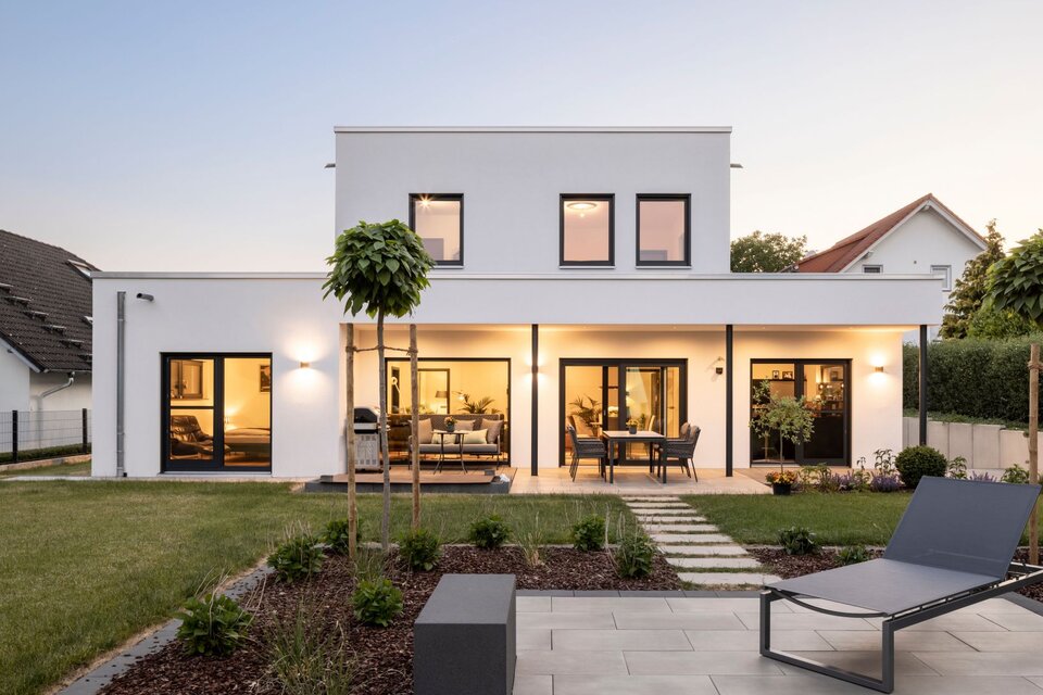 Abendliche Außenansicht eines frei geplanten FingerHaus im Bauhaus-Stil mit beleuchteter Terrasse und großen Fensterfronten. Im Vordergrund Garten mit moderner Liege und angelegten Beeten.