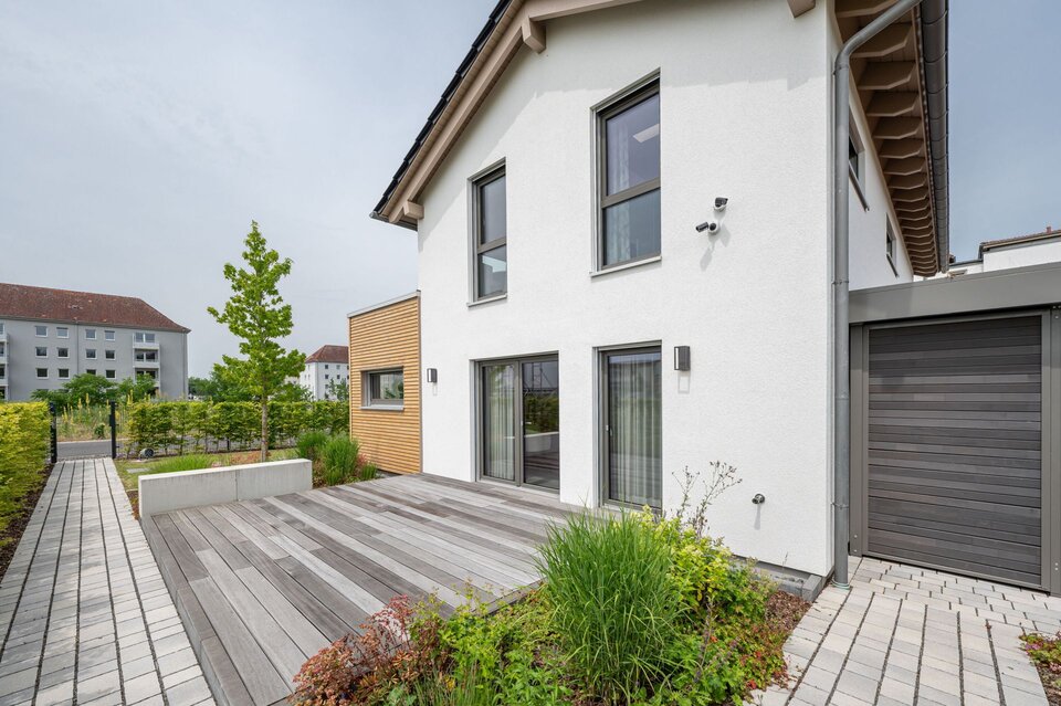 Außenbereich am Finger-Haus Musterhaus TALO in Babenhausen mit Holzterrasse, weißer Putzfassade, bodentiefen Fenstern, Holzfassaden-Element, gepflastertem Weg, bepflanzten Beeten und angrenzender Garage mit grauem Garagentor.