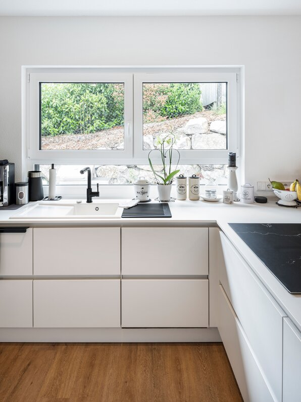 Küche des Kundenhauses NIVO 80 mit weißer Einbauküche, Spüle, Kochfeld und Fenster über der Arbeitsfläche.