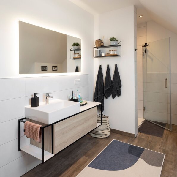Modernes Badezimmer mit bodengleicher Dusche, Waschtisch und großem Spiegel mit Beleuchtung. Holzoptikboden, helle Fliesen und schwarze Handtücher betonen das elegante Design.