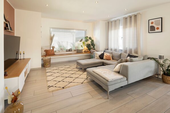 Wohnzimmer im FingerHaus SENTO 403 mit Ecksofa, Fensterbank, Teppich, Fernseher und bodentiefen Vorhängen